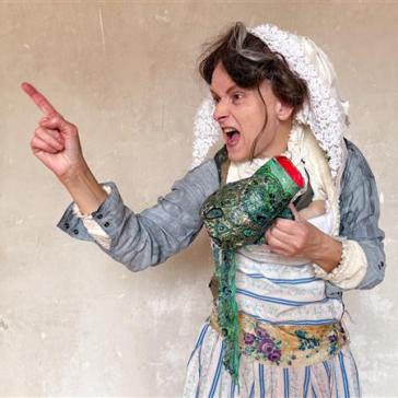 Person in historisch-verspielter Kleidung mit Haube zeigt energisch mit dem Finger nach links. In der anderen Hand hält sie eine bunt verzierte Handpuppe oder Requisite.
