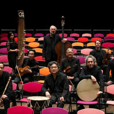 Zehn Musiker in schwarzer Kleidung posieren mit traditionellen und modernen Instrumenten. Sie sitzen und stehen zwischen bunten Stühlen auf einer Bühne. Die Atmosphäre wirkt professionell und kulturell.