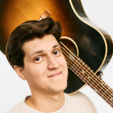 Ein junger Mann mit braunen, gepflegten Haaren und einem fröhlichen Lächeln trägt ein helles T-Shirt und hält eine Gitarre auf der Schulter. Er wirkt entspannt und freundlich.