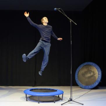 Ein Mann springt auf einem kleinen blauen Trampolin in die Luft, hebt einen Arm und steht vor einem Mikrofon auf der Bühne. Im Hintergrund sind ein weiteres Trampolin und ein Keyboard zu sehen.
