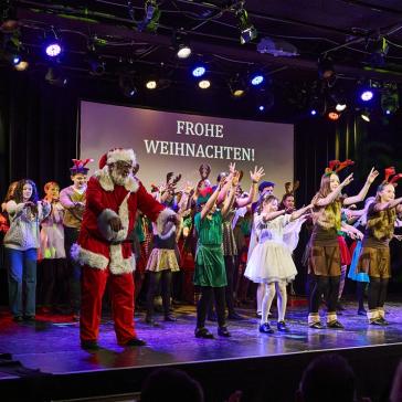 Eine große Gruppe verkleideter Menschen, darunter ein Weihnachtsmann, Elfen und Rentiere, steht auf einer festlich beleuchteten Bühne. Im Hintergrund leuchtet die Botschaft „Frohe Weihnachten!“.