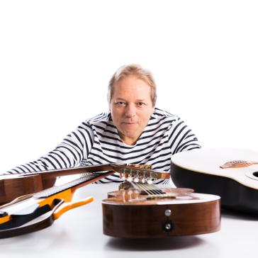 Ein Mann im gestreiften Shirt lehnt nach vorn und schaut in die Kamera. Vor ihm liegen mehrere Gitarren auf einem weißen Tisch, die er mit ausgestreckten Armen umfasst.