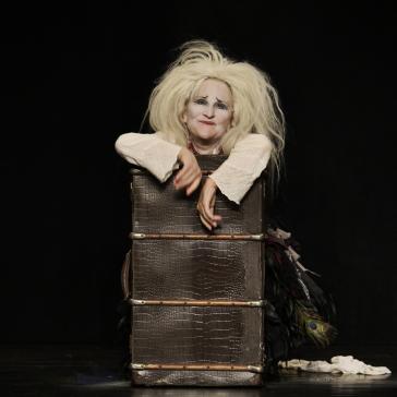 Eine Frau mit zerzausten, weißen Haaren und weiß geschminktem Gesicht lehnt in einem alten Koffer. Sie trägt ein weißes Oberteil und wirkt wie in einer theatralischen Szene.