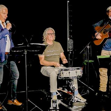 Drei Musiker auf der Bühne. Links mit Notenständer und Gesangsmikrofon, in der Mitte mit einer Snare-Drum und auf der rechten Seite mit einer Gitarre.