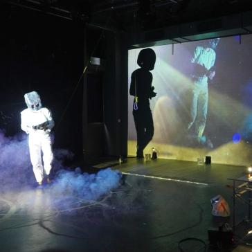 Eine Person im weißen Anzug mit Helm steht in Nebel, während rechts jemand an einem Projektortisch arbeitet. Im Hintergrund sind Projektionen von Figuren auf einer großen Leinwand zu sehen.
