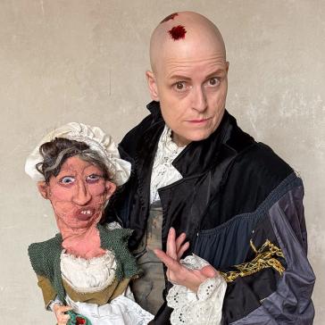 Person mit kahlem Kopf in historischer Robe posiert neben einer Puppenfigur, die wie eine ältere Frau mit Haube und Schürze aussieht.