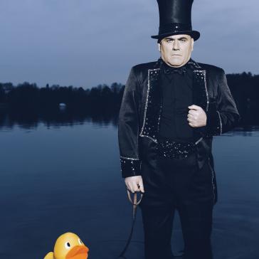 Ein Mann in schwarzem Frack und hohem Zylinder steht ernst im Wasser. An einer Leine hält er eine große gelbe Plastikente, die neben ihm im Wasser schwimmt.
