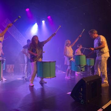 Eine Gruppe von Musikerinnen und Musikern spielt auf großen grünen Trommeln. Sie tragen helle Kleidung und heben die Schlägel rhythmisch in die Luft. Buntes Bühnenlicht erleuchtet die Szene.
