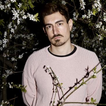 Mann mit ernstem Blick und hellem Strickpulli steht nachts vor einem blühenden Baum mit weißen Blüten und grünen Blättern.