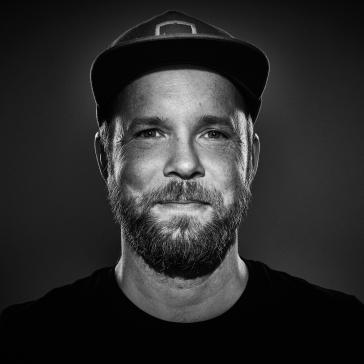 Porträt eines Mannes mit Basecap, Vollbart und ernstem Blick, fotografiert in Schwarz-Weiß mit dramatischer Beleuchtung.