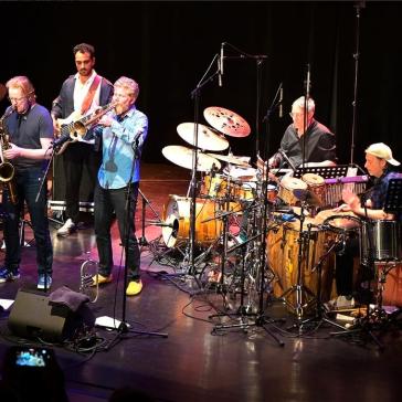 Fünf Musiker auf einer Bühne: Ein Pianist, ein Saxophonist, ein Trompeter, ein Schlagzeuger und ein Percussionist spielen vor Publikum.