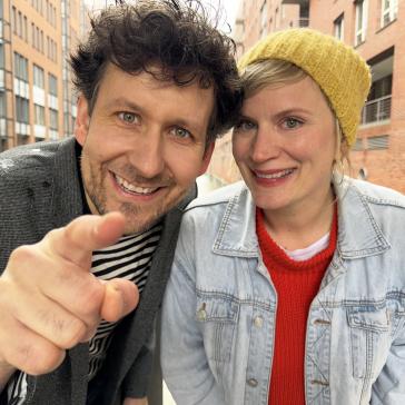 Zwei lächelnde Personen – eine mit gelber Strickmütze und Denim-Jacke, die andere mit dunklem Jackett über gestreiftem Shirt – zeigen auf etwas, im Hintergrund moderne Gebäude.