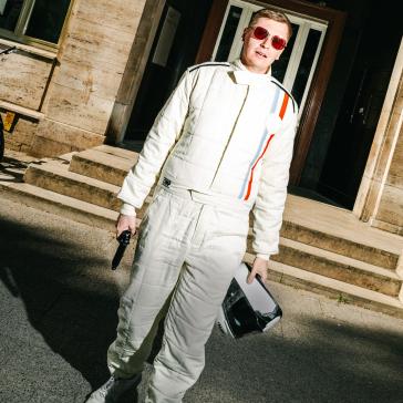 Person in einem weißen Rennfahreranzug mit roten Sonnenbrillen und Helm in der Hand steht auf einer Straße vor einem Gebäude mit Treppen.