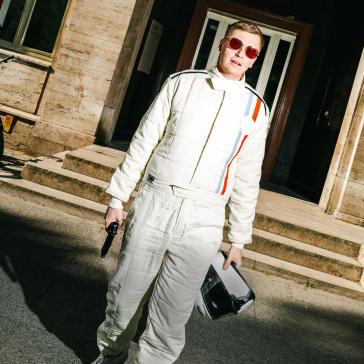 Person in einem weißen Rennfahreranzug mit roten Sonnenbrillen und Helm in der Hand steht auf einer Straße vor einem Gebäude mit Treppen.