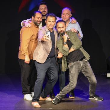 Fünf Männer posieren fröhlich auf einer Bühne: Einer macht ein Selfie, ein anderer hält ein Mikrofon, alle umarmen sich in blauer und roter Bühnenbeleuchtung.