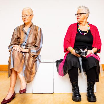 Zwei ältere Frauen sitzen nebeneinander: Eine trägt ein gestreiftes Kleid und Brille, die andere hält eine Kamera, trägt schwarze Kleidung, bunte Kette und robuste Stiefel.