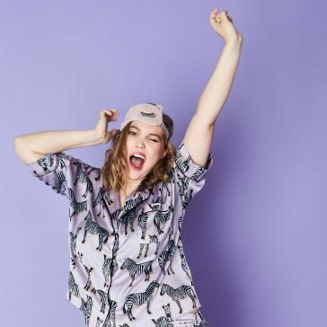 Jubelnde Person mit Schlafmaske auf dem Kopf, streckt die Arme nach oben, trägt einen lila Pyjama mit schwarzem Zebramuster und wirkt voller Freude.