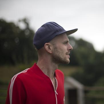 Mann mit blauer Basecap und rotem Sportshirt mit weißen Streifen steht im Profil, blickt nachdenklich in die Ferne, im Hintergrund verschwommene Natur mit Bäumen.
