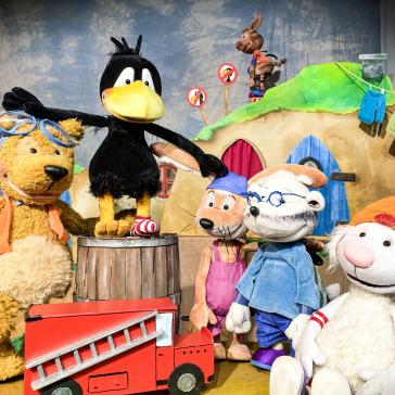 Fünf Stofftiere auf einer Bühne: Ein Bär mit Schwimmbrille, ein Rabe auf einem Fass, ein Kaninchen mit Latzhose, ein Schaf mit Brille und ein Lamm mit Mütze, vor einem Haus und Feuerwehrauto.