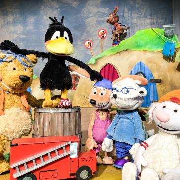 Fünf Stofftiere auf einer Bühne: Ein Bär mit Schwimmbrille, ein Rabe auf einem Fass, ein Kaninchen mit Latzhose, ein Schaf mit Brille und ein Lamm mit Mütze, vor einem Haus und Feuerwehrauto.