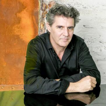 Mann mit graumelierten Haaren und schwarzem Hemd sitzt nachdenklich auf einem glatten Tisch vor einer rostigen Metallwand mit abblätternder Farbe.