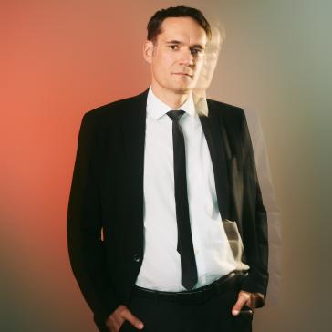 Mann im dunklen Anzug mit weißem Hemd und Krawatte steht mit verschränkten Händen vor einem farbigen, grün-rötlichen Hintergrund und wirkt nachdenklich.