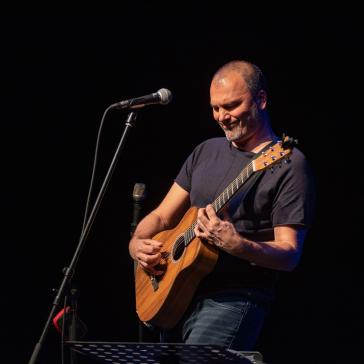 Mann mit Gitarre und dunklem T-Shirt spielt konzentriert auf einer Bühne, ein Mikrofonstativ vor ihm, im Hintergrund dunkel – entspannte, musikalische Atmosphäre.
