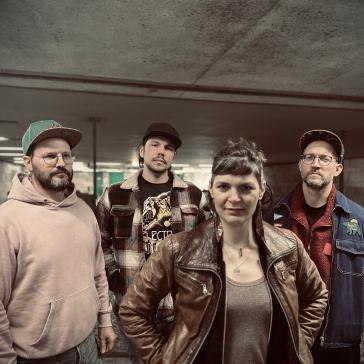 Vier Personen in lässiger Kleidung – zwei mit Basecaps, eine mit Lederjacke, eine mit rosafarbenem Hoodie – posieren in einem Tunnel mit Betonwänden und Neonlicht.