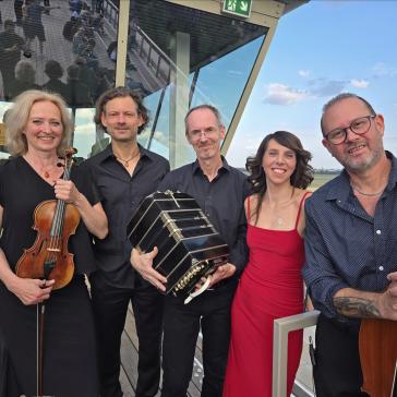 Fünf lächelnde Musiker: Eine Frau mit Geige, ein Mann mit Akkordeon, eine Frau in rotem Kleid, und zwei Männer mit Kontrabass, posieren auf einer Aussichtsplattform mit Glasbrüstung.