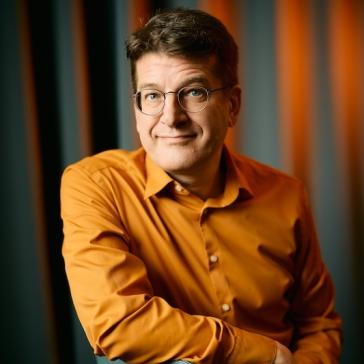 Lächelnder Mann mit Brille und orangefarbenem Hemd lehnt entspannt mit verschränkten Armen auf einem blauen Stuhl vor einem dunklen, vertikal gestreiften Hintergrund.