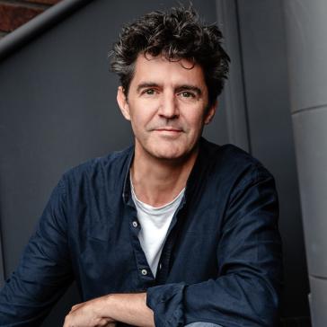 Mann mit struppigen Haaren, dunklem Hemd und Jeans sitzt entspannt vor einer grauen Wand mit Treppengeländer, die Arme lässig auf den Knien abgelegt.