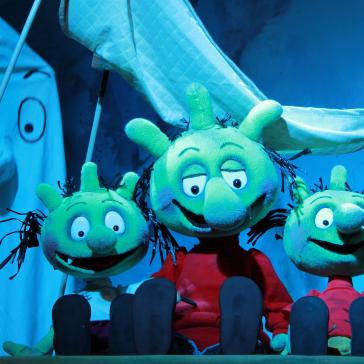 Drei grüne, lächelnde Puppenfiguren mit strubbeligen Haaren sitzen nebeneinander auf einer Bühne, im Hintergrund ein weißes Gespenst mit Stab und eine schwebende Banane.