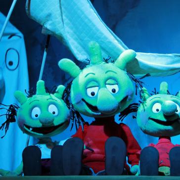 Drei grüne, lächelnde Puppenfiguren mit strubbeligen Haaren sitzen nebeneinander auf einer Bühne, im Hintergrund ein weißes Gespenst mit Stab und eine schwebende Banane.