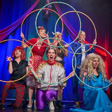 Sechs bunt gekleidete Schauspieler posieren mit Reifen auf einer Bühne: Kostüme in Rot, Blau, Lila, Grau und Perücken, vor blauem Vorhang mit roter Drapierung.