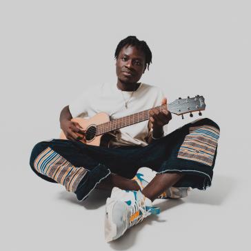 Junger Mann sitzt im Schneidersitz, spielt Gitarre, trägt ein weißes T-Shirt, bunte Hosen mit Streifen und weiße Sneaker mit orange-blauen Akzenten, vor neutralem Hintergrund.