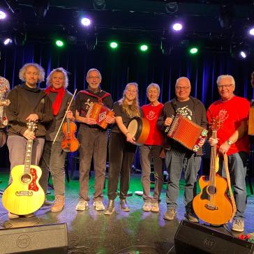 Acht lächelnde Musiker auf einer Bühne: Mit Gitarren, Geige, Akkordeons, Banjo und Trommel, bunt gekleidet, vor blau beleuchtetem Bühnenhintergrund.