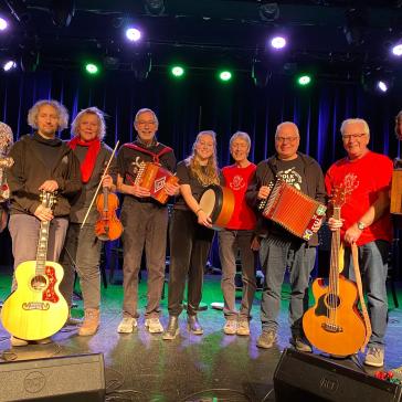 Acht lächelnde Musiker auf einer Bühne: Mit Gitarren, Geige, Akkordeons, Banjo und Trommel, bunt gekleidet, vor blau beleuchtetem Bühnenhintergrund.
