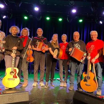 Acht lächelnde Musiker auf einer Bühne: Mit Gitarren, Geige, Akkordeons, Banjo und Trommel, bunt gekleidet, vor blau beleuchtetem Bühnenhintergrund.