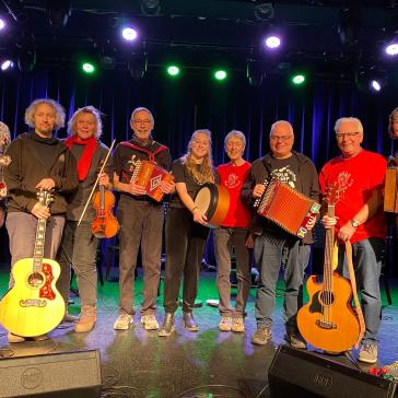 Acht lächelnde Musiker auf einer Bühne: Mit Gitarren, Geige, Akkordeons, Banjo und Trommel, bunt gekleidet, vor blau beleuchtetem Bühnenhintergrund.