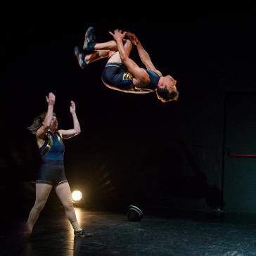 Zwei Akrobaten auf dunkler Bühne: Eine Frau steht mit erhobenen Armen, ein Mann fliegt kopfüber durch die Luft, im Hintergrund eine Person mit Kleiderständer und Seil.