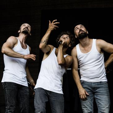 Drei Männer in weißen Unterhemden und Jeans: Der linke streckt Arm aus, der mittlere singt in ein Mikrofon, der rechte lehnt sich lässig zurück, alle vor schwarzem Hintergrund.