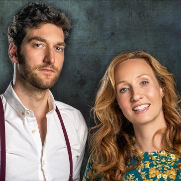 Zwei lächelnde Personen vor metallisch-blauem Hintergrund: Mann mit weißem Hemd und Hosenträgern, Frau mit langem rotblondem Haar in blumigem Oberteil.