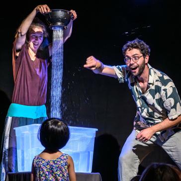 Bühnenbild: Frau gießt Wasser aus einem Sieb in eine durchsichtige Kiste, vor der ein Kind steht; rechts kniet ein lächelnder Mann in Hemd und lässiger Hose.