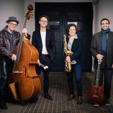 Vier lächelnde Musiker vor weiß-grauer Ziegelwand: Mann mit Kontrabass und Hut, Mann im Anzug, Frau mit Saxophon, Mann mit E-Gitarre und Schal.
