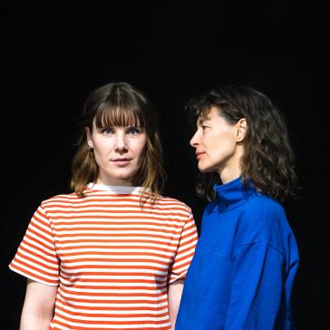 Zwei Frauen vor schwarzem Hintergrund: Eine mit rot-weiß gestreiftem T-Shirt und Pony, eine mit dunkelblauem Rollkragenpullover und lockigem Haar, beide leicht seitlich zueinander stehend.