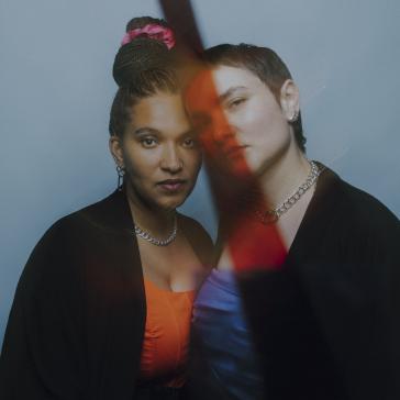 Zwei Personen eng beieinander: Eine mit hochgesteckten Haaren, pinkem Haarreif und silberner Kette, eine mit kurzem Haar und Kette, vor blauem Hintergrund mit farbigem Lichteffekt.