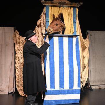 Mann in schwarzem Mantel und Zylinder füttert ein Pferd, das aus einer blauen Krippe mit weißen Streifen lugt, auf einer Bühne mit beigefarbenen Vorhängen.