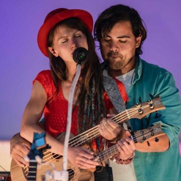 Zwei Musiker auf der Bühne: Frau mit rotem Hut singt ins Mikrofon, Mann mit Dreadlocks spielt Ukulele, beide konzentriert und nah beieinander.