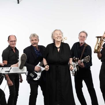 Sechs lächelnde Musiker: Frau am Keyboard, Mann mit hochgestelltem Fuß, zwei Gitarristen, Sängerin in schwarzem Kleid, Saxophonist – alle in schwarzer Kleidung vor weißem Hintergrund.