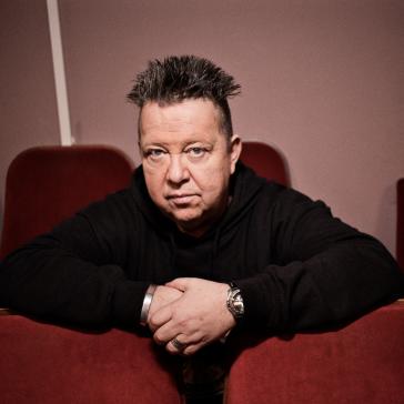 Mann mit aufrecht stehendem Haar und schwarzem Pullover sitzt nachdenklich mit verschränkten Armen auf roten Kinosesseln, Blick direkt in die Kamera.